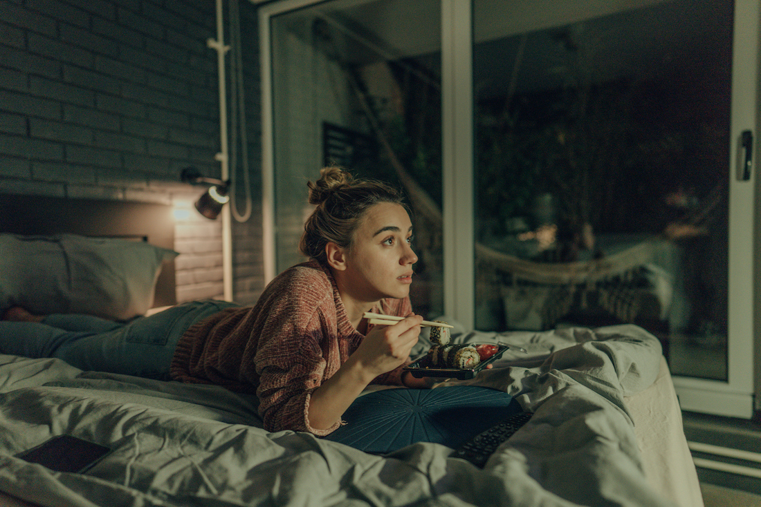 Photo of a young woman watching TV in the bedroom of her apartment; eating sushi and enjoying her night at home alone.
